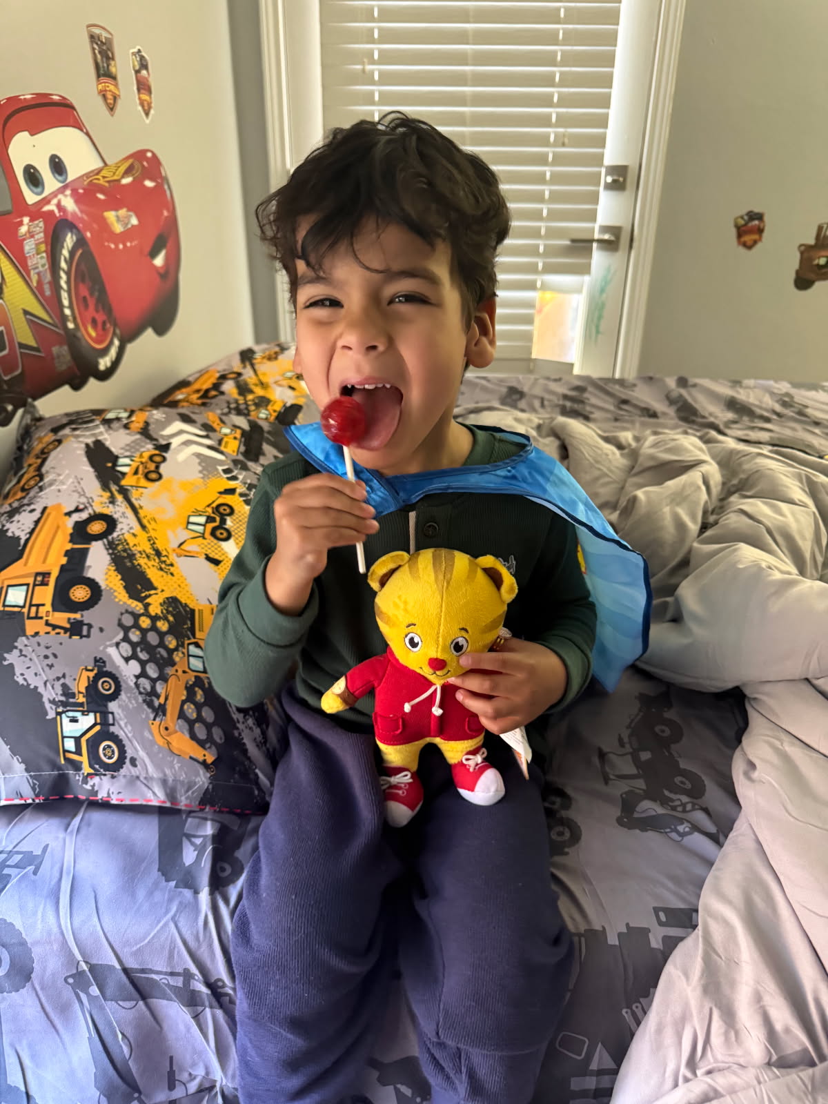Young boy holds a red lollipop and a stuffed Daniel Tiger on his bed with yellow dump truck linens.